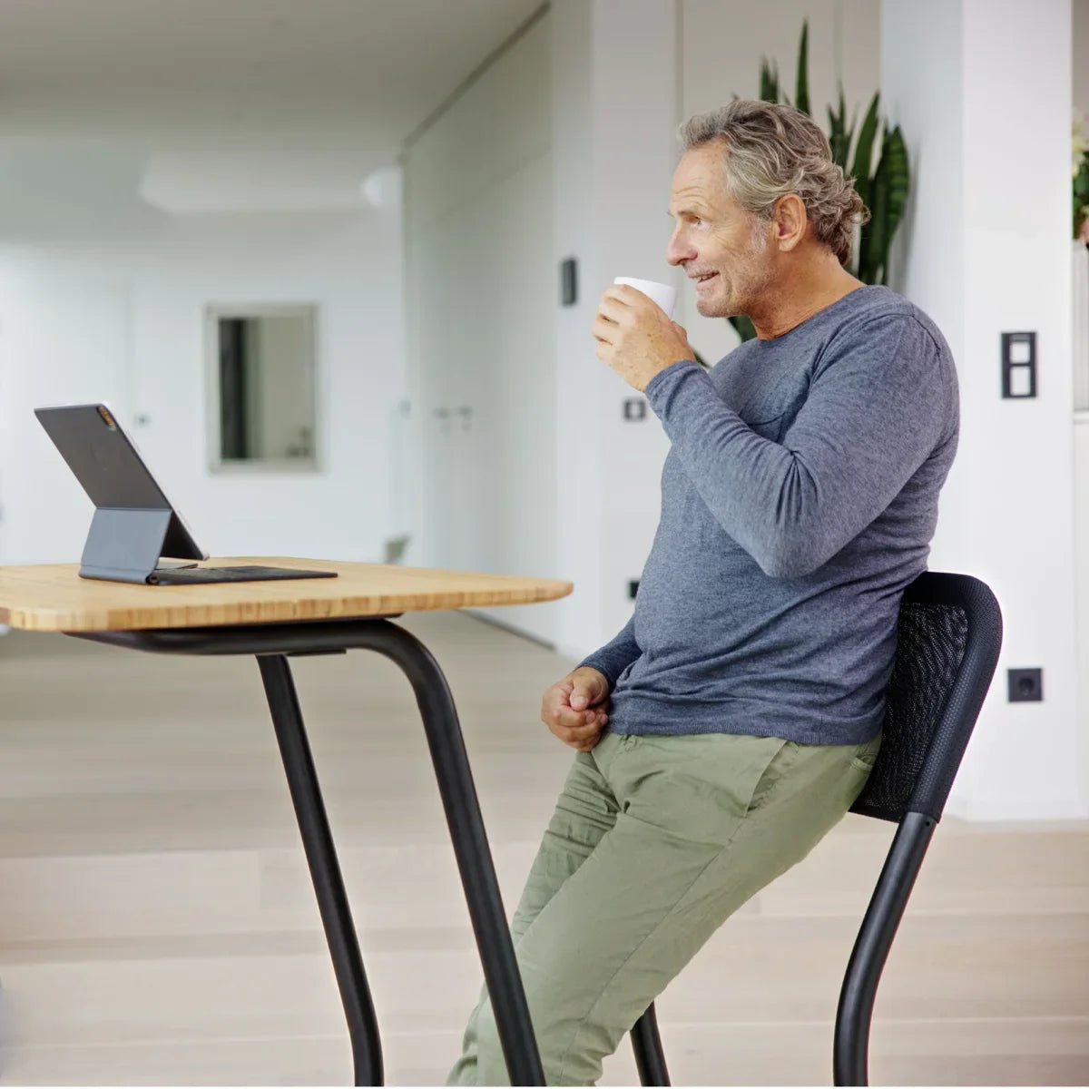 A middle-aged man sits at a tall table, drinking from a cup and smiling while looking at a tablet on a stand in a bright, modern room with light wood floors.