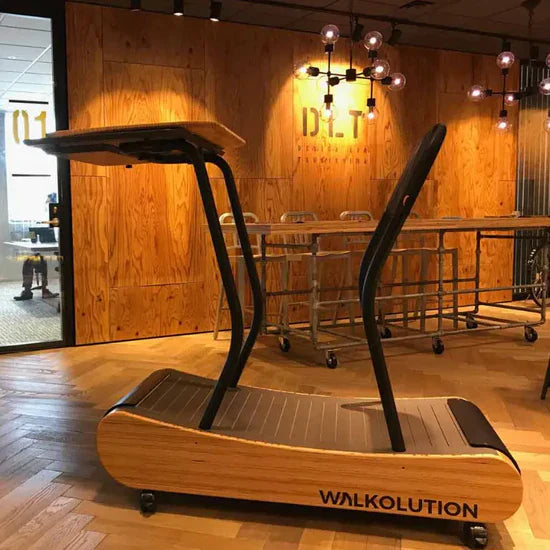 A wooden treadmill with a built-in desk, branded WALKOLUTION, is positioned in a modern office space with wooden walls, industrial lighting, and metal chairs.