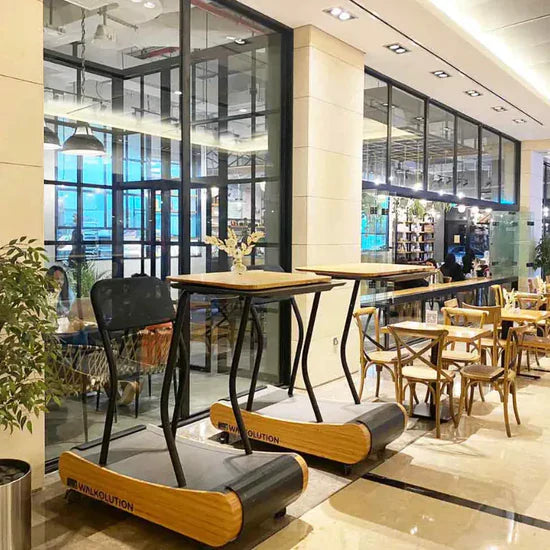 Two treadmills with attached wooden desks are placed in a modern, well-lit cafe or coworking space with glass walls, wooden tables, chairs, and plants. People can work while walking on the treadmills.