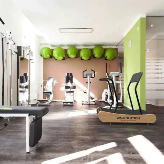 A bright, modern gym with exercise equipment, including a treadmill, weight machines, and a row of green stability balls mounted high on the back wall. Natural light fills the clean, organized space.