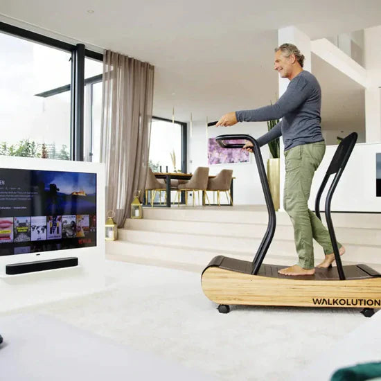 A person walks barefoot on a wooden treadmill in a modern, bright living room while watching TV. The treadmill is labeled WALKOLUTION, and the space features large windows and stylish furniture.
