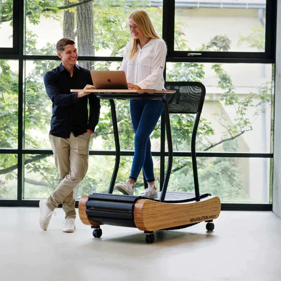A woman stands and works on a laptop at a desk that is built on top of a treadmill, while a man stands nearby smiling. They are in a bright room with large windows showing greenery outside.