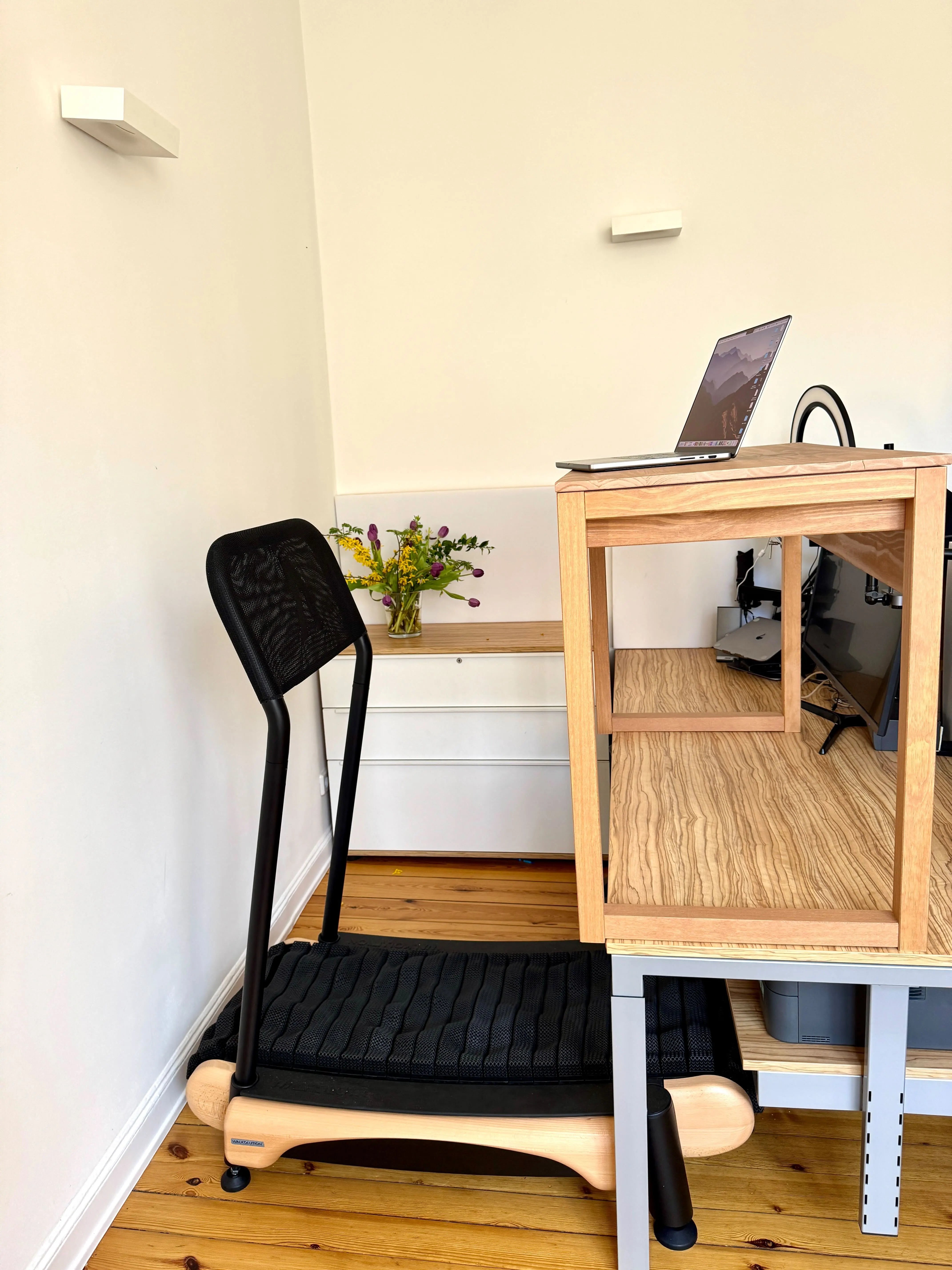 A modern home office setup with a treadmill desk, a laptop on a wooden desk, a black treadmill chair, a drawer unit, and a vase of flowers against a white wall on a wooden floor.