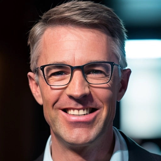 A smiling man with short, light brown hair and glasses, wearing a dark blazer and a light-colored shirt, stands indoors with blurred lights in the background.