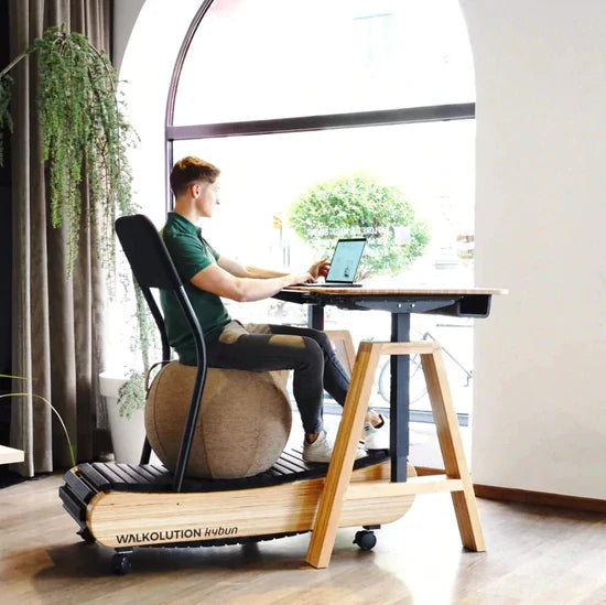 A man works on a laptop at a standing desk, seated on a stability ball placed on a wooden treadmill workstation, in a bright room with large arched window and plants.