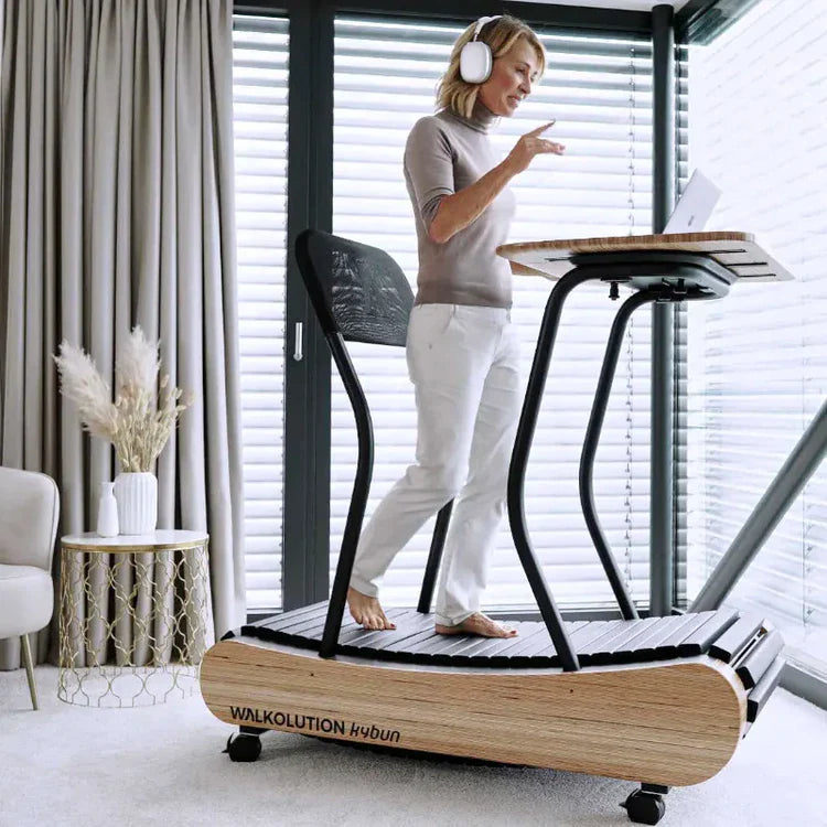 A woman wearing headphones walks barefoot on a wooden treadmill desk while working on a laptop in a bright, modern room with large windows and light-colored decor.