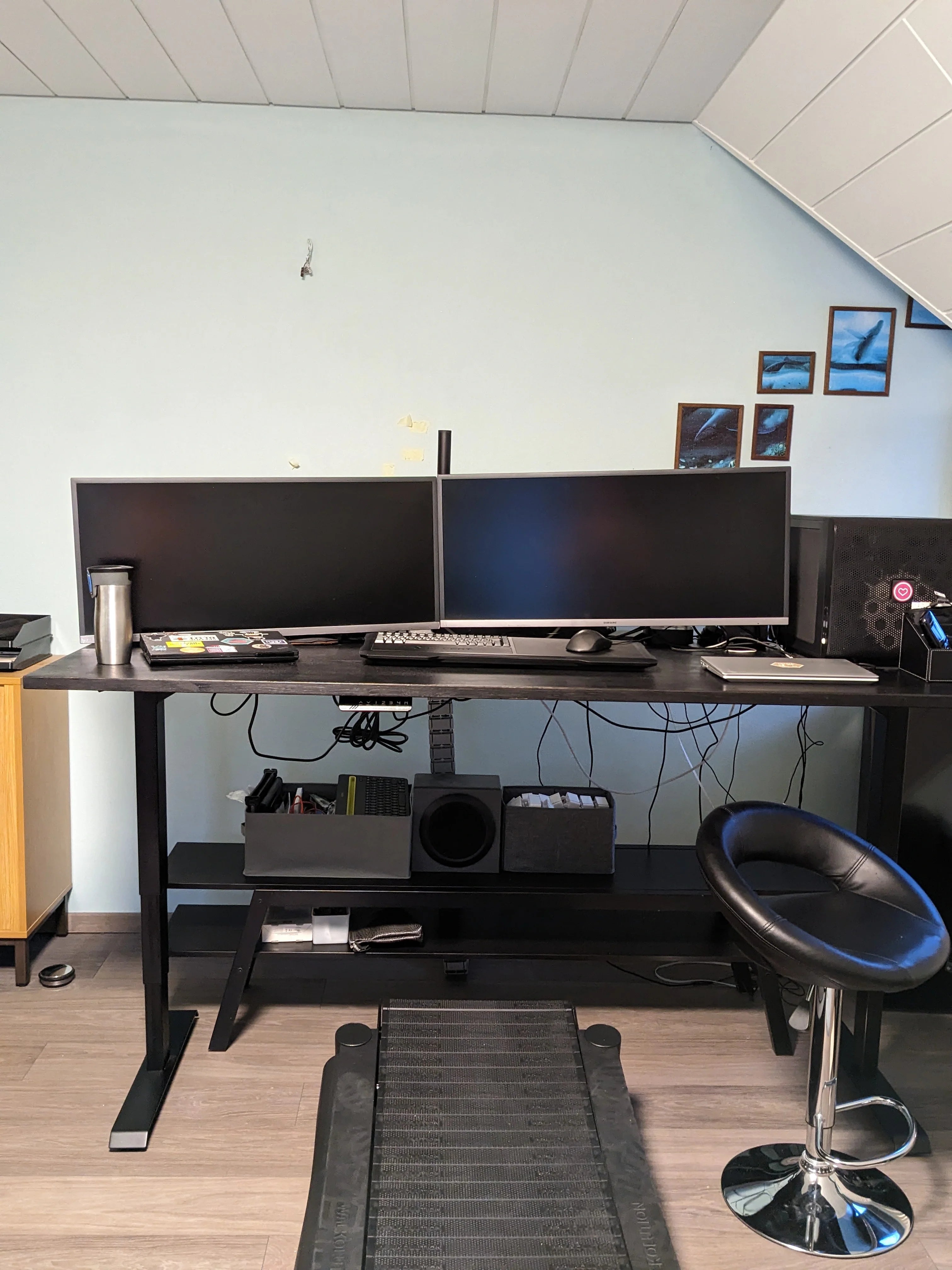 A home office setup with two large monitors on a black standing desk, a keyboard, mouse, and accessories. A black swivel chair and treadmill are in front, with pictures and speakers on the light blue wall behind.