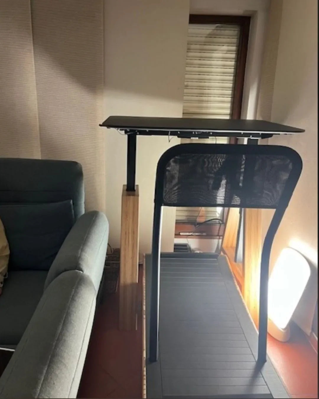 A treadmill with a black mesh back and a raised desk board in front stands next to a teal sofa, near a window with blinds and a bright floor lamp.
