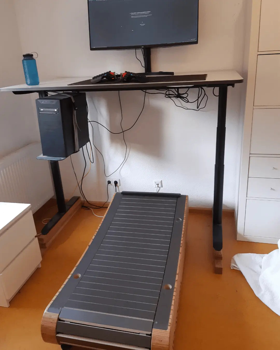 A standing desk setup with a computer monitor, keyboard, and water bottle on top, and a treadmill below for walking while working. Cables hang under the desk, and a white cabinet is nearby.