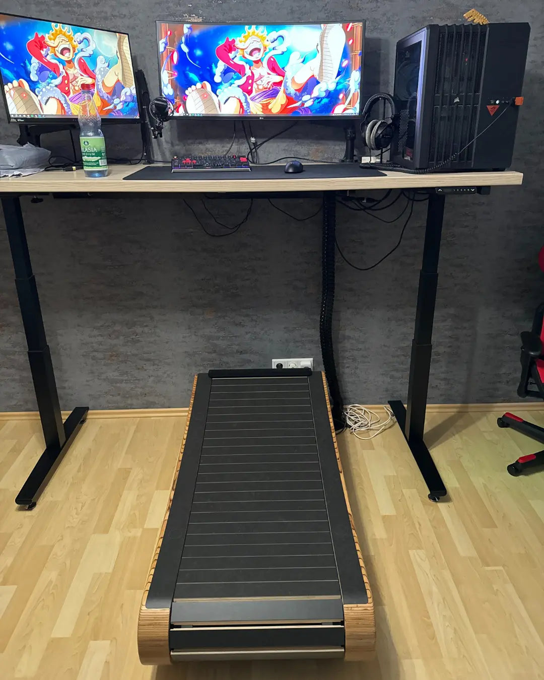 A standing desk with a computer setup, dual monitors displaying an anime character, and a treadmill placed underneath, allowing for walking while working. Cables and a water bottle are also visible on the desk.