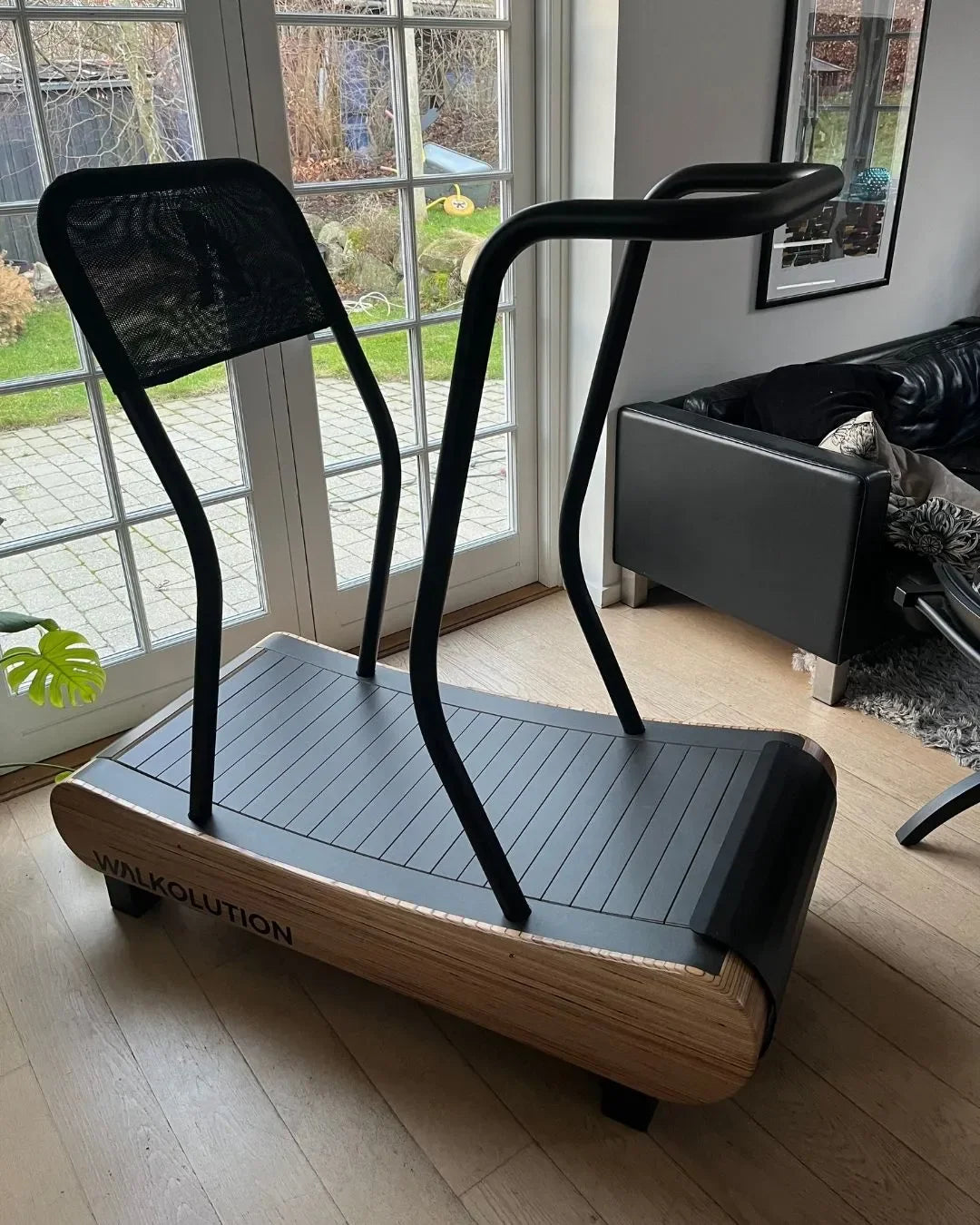 A modern, curved, non-motorized wooden treadmill with black handles sits in a bright living room near large windows and a black sofa. The treadmill is labeled WALKOLUTION.