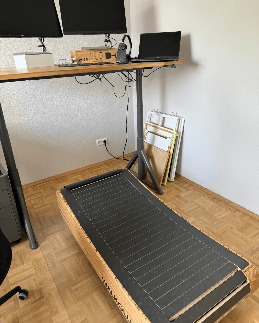 A standing desk with monitors, a laptop, and headphones is set up in a room with a wooden-tiled floor. In front of the desk is a compact treadmill, and some framed pictures are stacked against the wall.