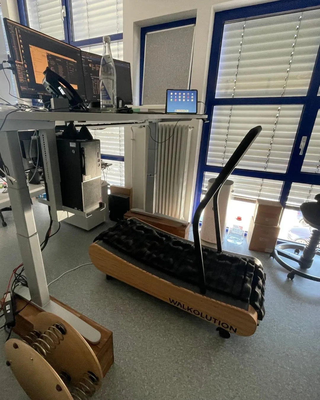 A modern office with a standing desk setup and a WALKOLUTION manual treadmill positioned beneath the desk, allowing the user to walk while working on the computer. Sunlight filters through blue-framed windows with blinds.