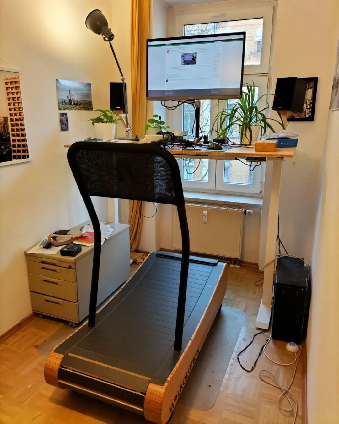 A treadmill is positioned in front of a standing desk with a large monitor, keyboard, plants, and other items on it. The setup is in a small, bright room with art on the walls and a window letting in natural light.
