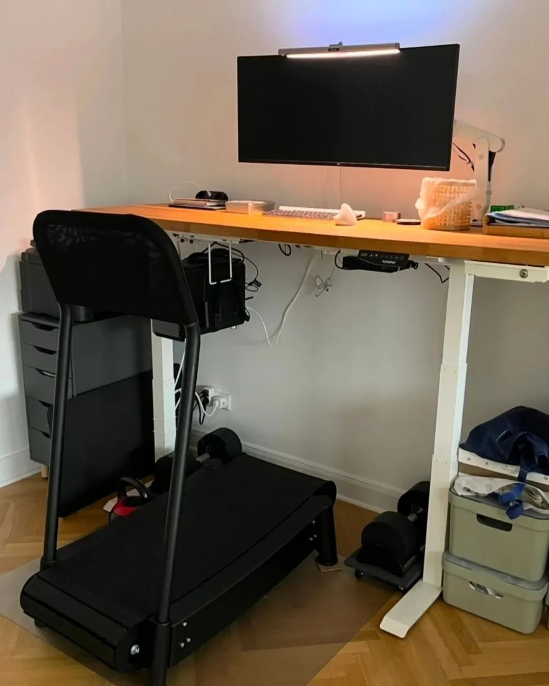 A home office setup with a large monitor, keyboard, and mouse on a wooden standing desk. A treadmill is positioned beneath the desk, allowing for walking while working. Various office supplies are organized nearby.