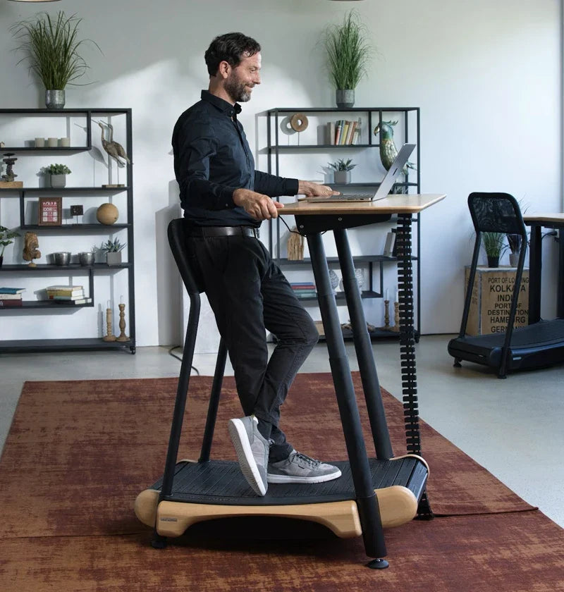 A man uses a standing desk with a built-in treadmill in a modern office, typing on a laptop. Bookshelves, decor, and plants are visible in the background.