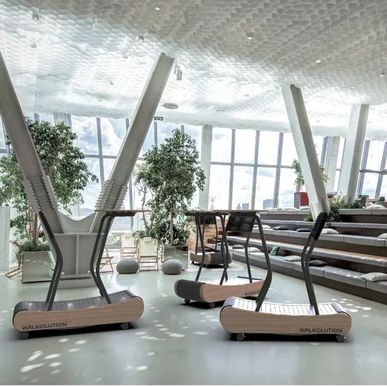 Two wooden manual treadmills labeled WALKOLUTION stand in a bright, modern indoor space with large windows, green plants, and tiered seating in the background. Light streams in, creating an airy atmosphere.