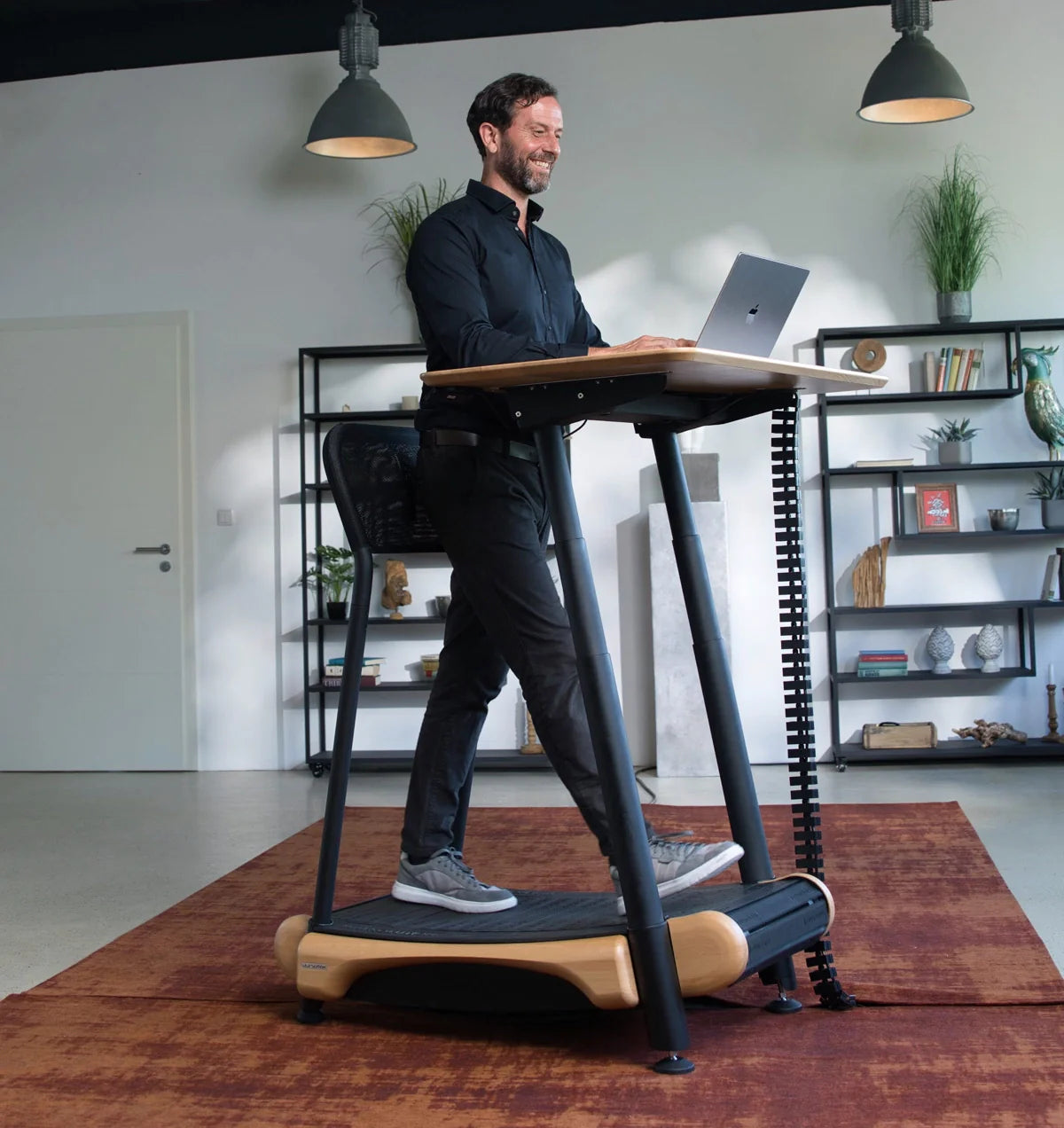 A man smiles while using the Walkolution Fusion Desk treadmill desk, working on a laptop at an electric height adjustable desk in a modern office with shelves, décor, and pendant lights in the background.