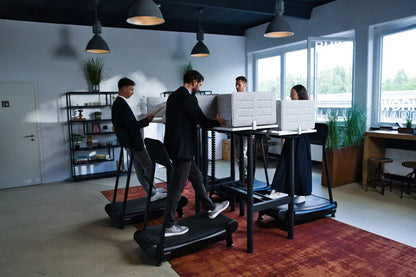 Four people stand at Walkolution Forum Conference Desks in a modern office with large windows, black shelving, plants, and overhead lamps. Each uses a computer, promoting an active work environment.