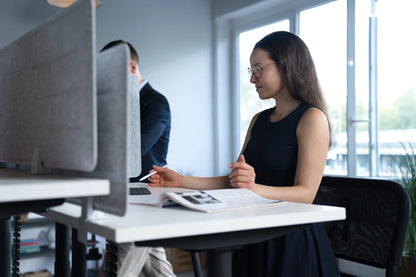 A woman in glasses and a black dress works at a Walkolution Forum Conference Desk with a magazine and laptop in a modern office. Another person is partially visible at an adjacent workstation behind the divider.