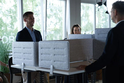 Three people stand around a Walkolution Forum Conference Desk with gray privacy dividers, smiling and chatting in a modern office filled with large windows and green plants.