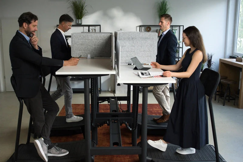 Four people use Walkolution Forum Conference Desks with privacy panels in a bright, modern office. Standing or leaning at height-adjustable desks with integrated treadmills, they work on laptops amid plants and natural light.