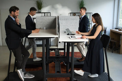 Four people use Walkolution Forum Conference Desks with privacy panels in a bright, modern office. Standing or leaning at height-adjustable desks with integrated treadmills, they work on laptops amid plants and natural light.