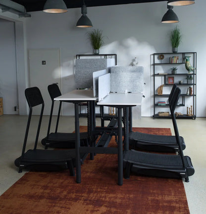 A modern office features four height-adjustable Walkolution Forum Conference Desks arranged around two white tables with gray privacy panels, while shelves with plants and decor stand in the background under pendant lights.
