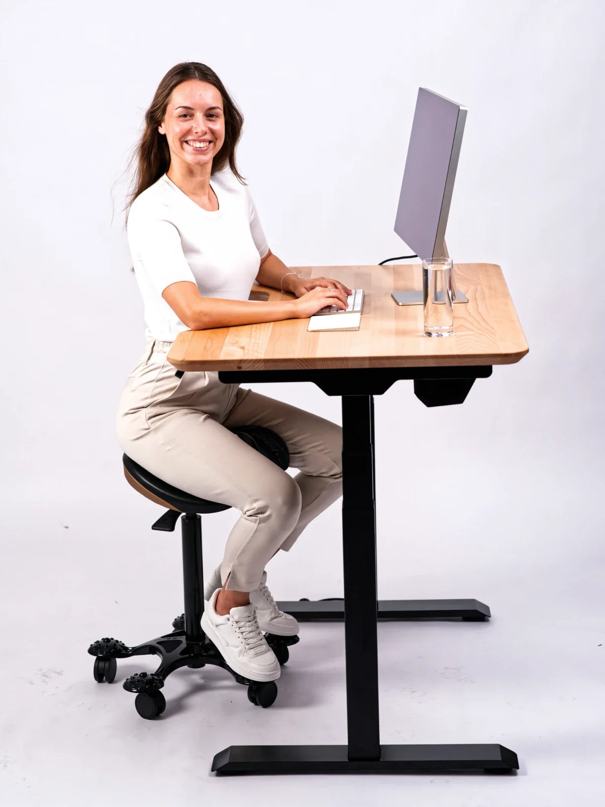 A woman smiles at the camera while sitting on an ergonomic stool at a Walkolution Core Desk made of solid beech wood. The desk is height-adjustable and holds a keyboard, computer monitor, and glass of water.