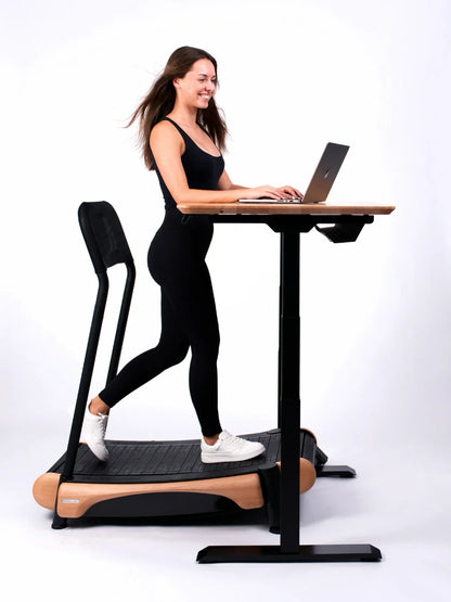 A woman in black activewear smiles while using the Walkolution Core Desk—a height-adjustable standing desk with a treadmill workstation—working on her laptop against a plain white background.