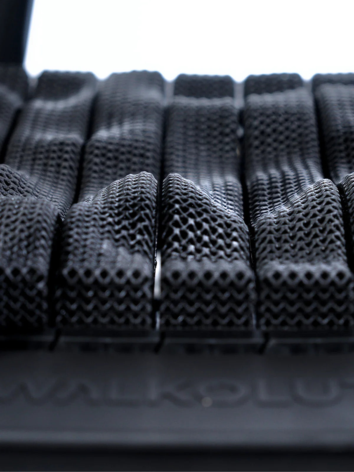 Close-up view of a black, textured surface with raised, mesh-like rectangular sections arranged in rows, resembling a flexible or adaptive cushioning material. Walkolution Global