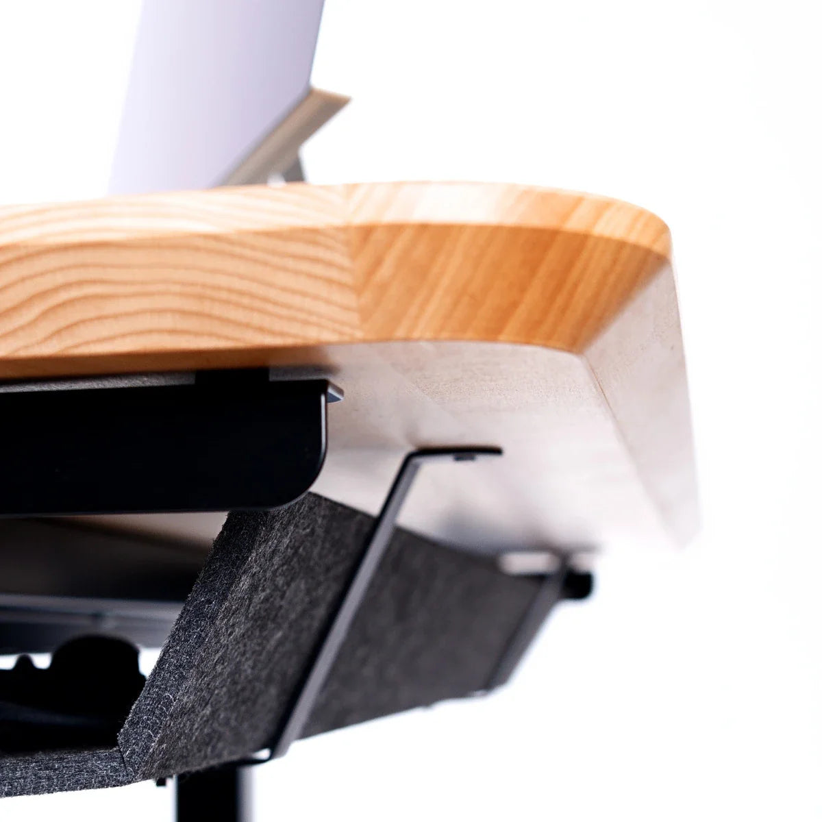 Close-up view of the underside of a wooden desk with rounded edges, showing a mounted gray felt storage compartment and part of a laptop on top. The background is white and uncluttered.