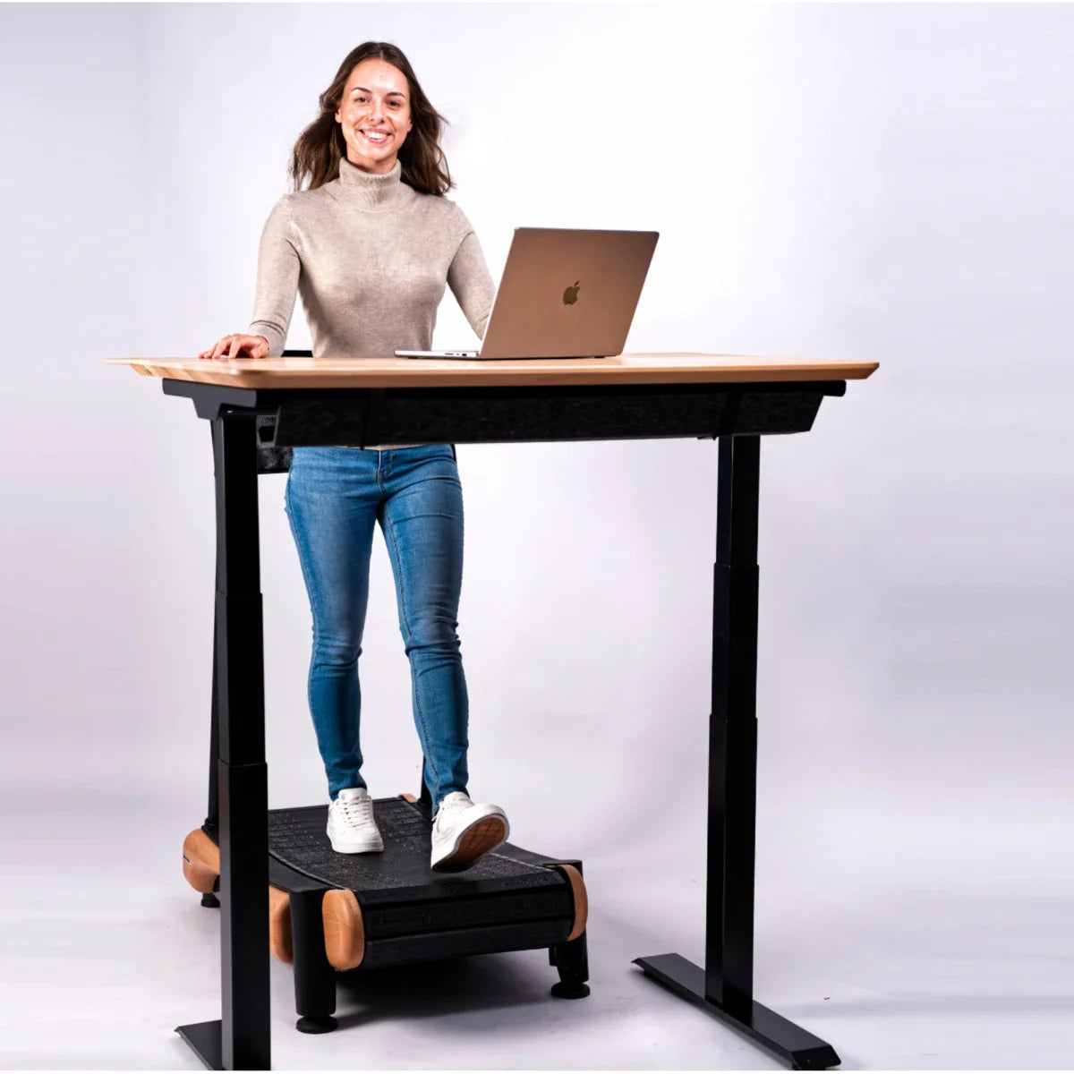 A woman stands at a height-adjustable desk using a laptop. She is smiling and walking on an under-desk treadmill. The background is plain white.