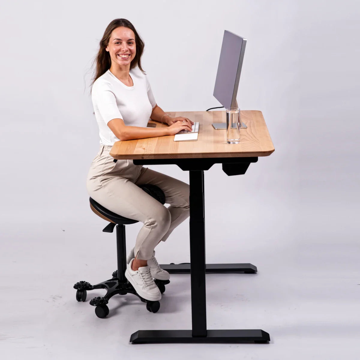A woman wearing a white shirt and beige pants sits on an ergonomic stool at a wooden standing desk, typing on a keyboard with a monitor and a glass of water in front of her, smiling at the camera.