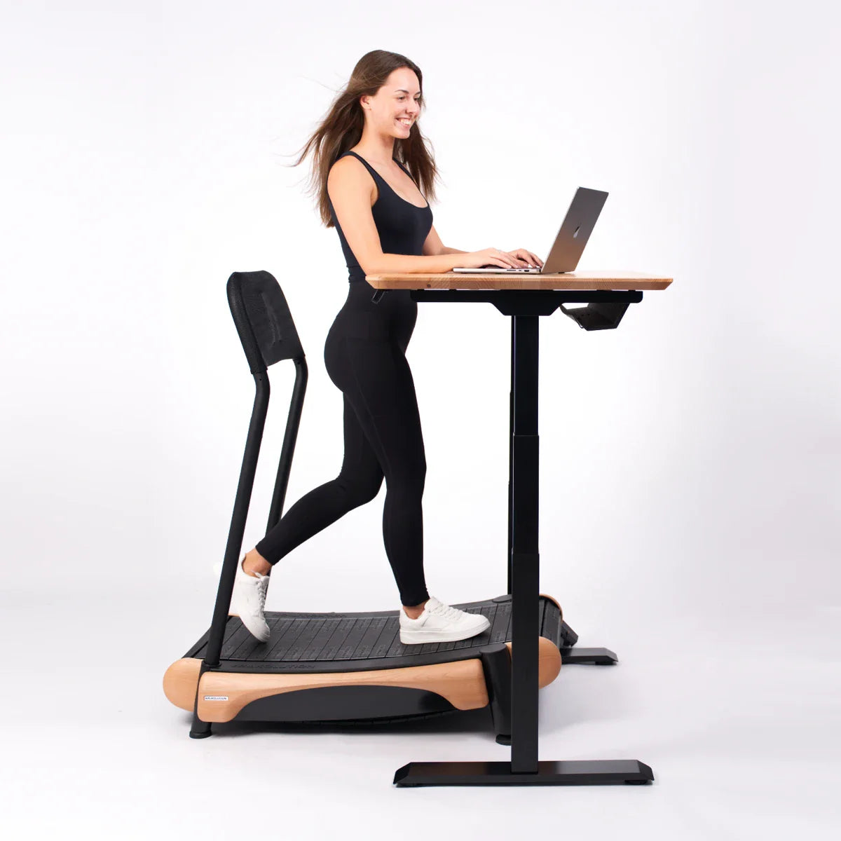 A woman in black activewear is smiling while walking on a treadmill and working on a laptop at a standing desk in a bright, minimalist setting.