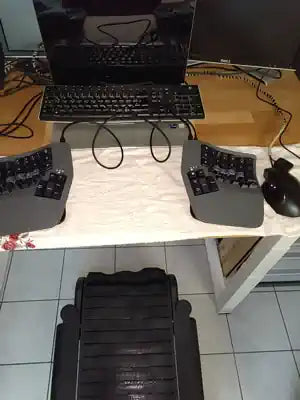 A computer setup with a monitor, a standard keyboard, a split ergonomic keyboard, a mouse, and a black foot pedal, all placed on a wooden desk above a tiled floor.