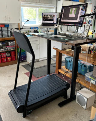 A standing desk with dual monitors, keyboard, and laptop is set up in a well-lit room. In front of the desk is a black treadmill, creating a walking workstation. Various tools and storage bins are visible in the background.