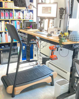 A home office with a standing desk setup, computer monitors, various electronics, and tools. A treadmill is positioned in front of the desk, allowing for walking while working. Bookshelves and organized cables are in the background.