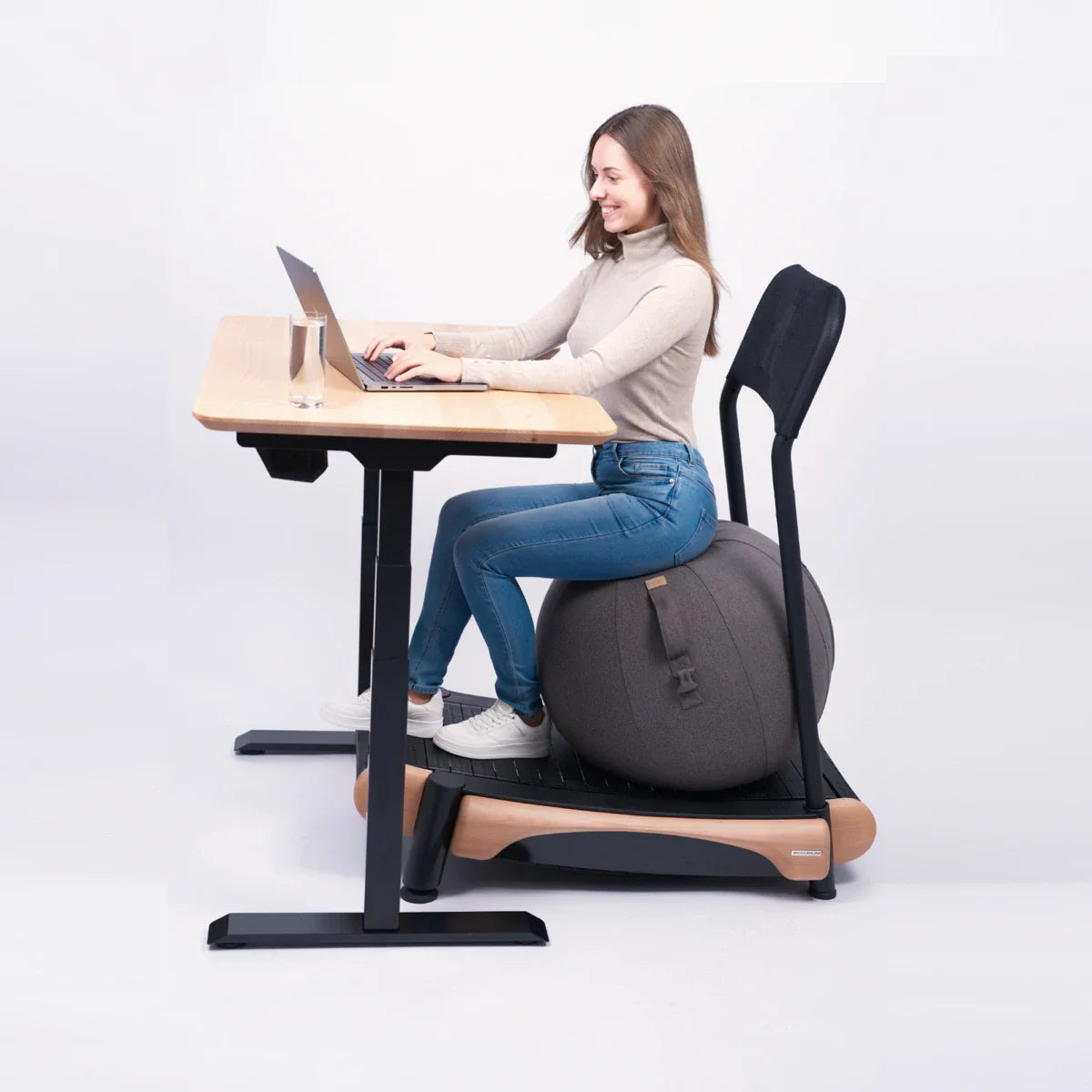 A woman smiles as she works at her laptop on a wooden desk, seated on a large gray Walkolution Active Sitting Ball with backrest. She's wearing a beige sweater and jeans against a plain white background.