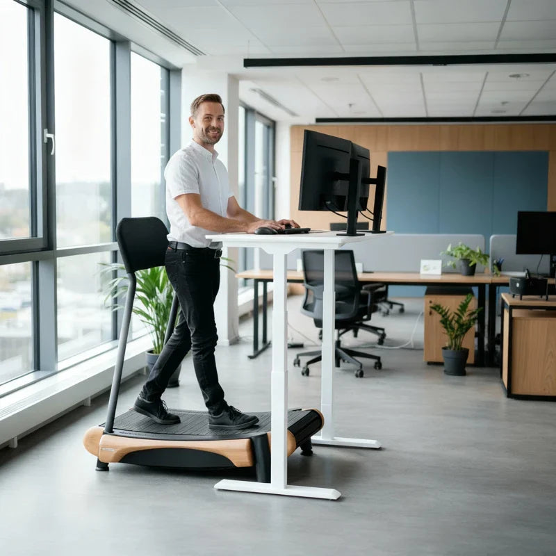 A man uses a treadmill desk while working on a computer in a modern office with large windows, plants, and several desks in the background.