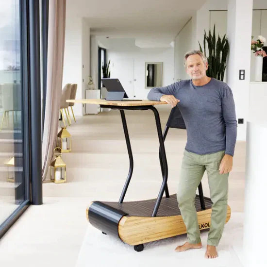 A man stands barefoot indoors, leaning on a modern wooden treadmill desk with a laptop on top. The space is bright, minimalistic, and features large windows, dining chairs, and decorative plants.