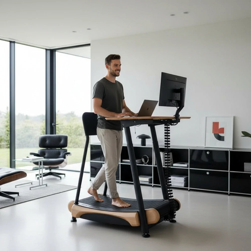 A man works on a laptop at a standing desk with a monitor, while walking barefoot on a treadmill in a modern, bright home office with large windows and minimalist décor. Walkolution Global