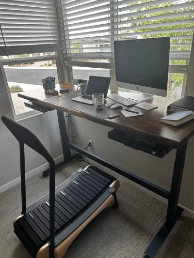 A modern home office setup with a treadmill desk, chair, computer monitor, laptop, mouse, phone, notebooks, pens, and a glass of iced coffee by a window with blinds partially open.