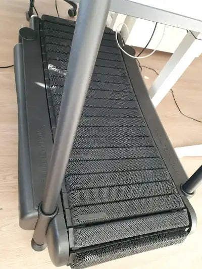 A compact treadmill placed under a desk, showing its black running belt and frame on a light wood floor; desk legs and electrical cords are visible above.