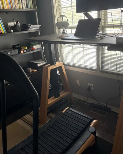 A home office with a treadmill under a standing desk, a laptop and monitor on the desk, books and office supplies on shelves, and large windows letting in natural light.