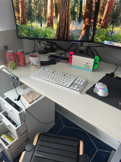 A white desk with a keyboard, mouse, two monitors displaying a forest scene, two soda cans, a mug, a Nintendo Switch, and various desk items. Drawers are open beneath the desk, and a chair is partially visible.