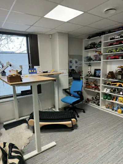 A modern office with a standing desk, a blue office chair, a treadmill under the desk, and shelves filled with model cars and toys. A window lets in natural light, and a white rug is on the floor.