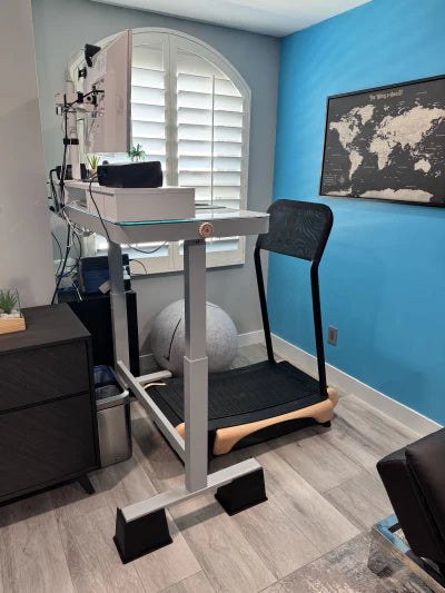 A modern home office setup featuring a standing desk positioned over a treadmill, with a computer monitor, keyboard, and mouse on the desk. The room has blue walls, a world map, and a window with white shutters.