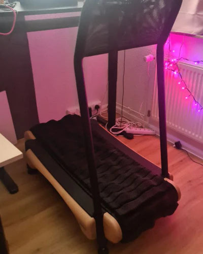 A compact treadmill stands in a room with pink LED string lights along a radiator. The treadmill has a black frame and textured running surface, and nearby desks and cables are visible on the wooden floor.