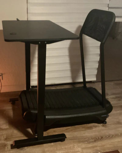 A compact treadmill desk with a black tabletop and mesh-back chair stands on a wooden floor in front of closed white blinds.